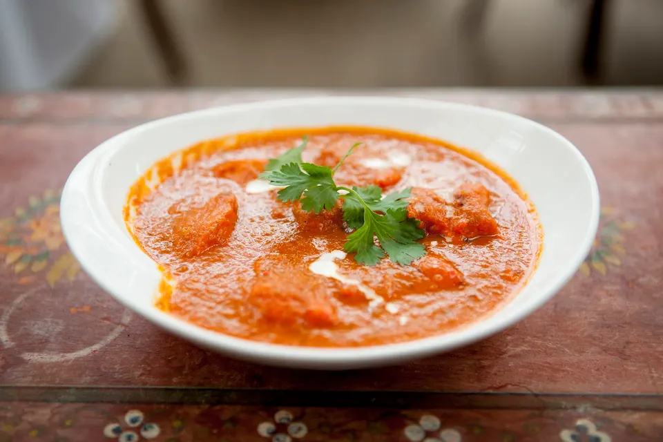 A white bowl filled with creamy orange chicken tikka masala curry, garnished with fresh cilantro leaves and a swirl of cream, served on a rustic wooden table.