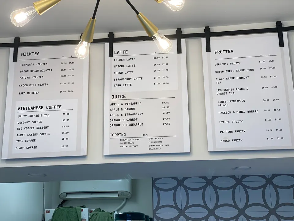 Three white menu boards hanging from a black metal rail at Lermer Cafe, displaying options for milk tea, Vietnamese coffee, lattes, juices, toppings, and fruit teas with prices.