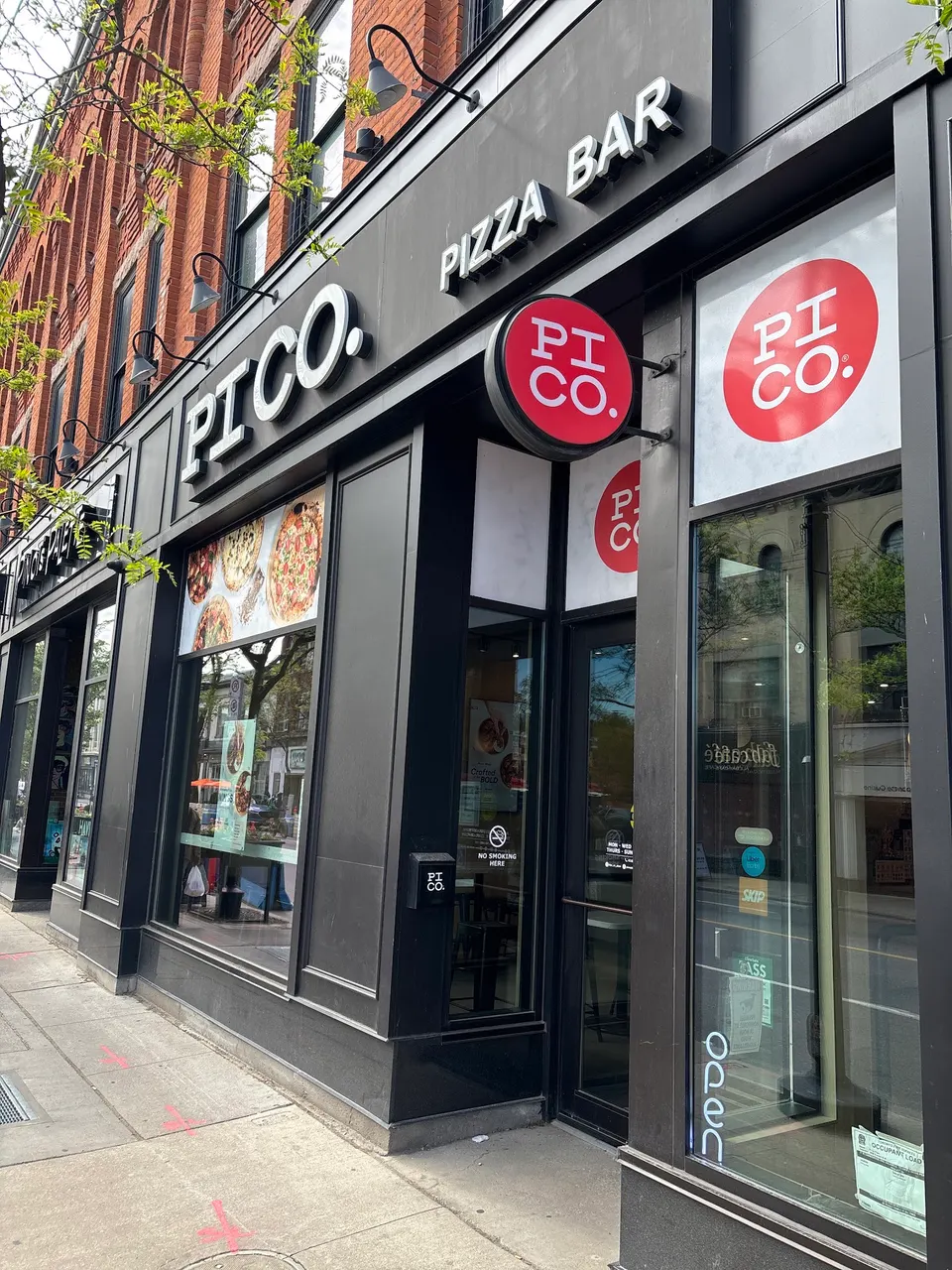 Exterior storefront of Pi Co. Pizza Bar featuring a black facade, large windows, and red circular logos on the glass and hanging sign.