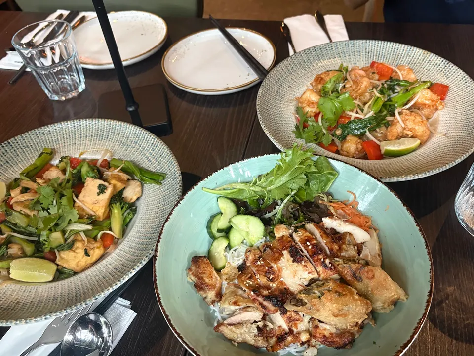 Three Vietnamese dishes served on a dark wooden table, including a chicken and spring roll vermicelli bowl, a stir-fried tofu dish, and a prawn stir-fry with vegetables.