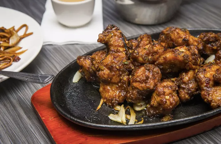 A sizzling hot plate piled high with crispy, glazed chicken pieces garnished with onions, served on a wooden trivet alongside other Chinese dishes.