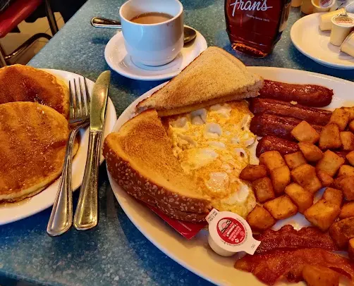 A hearty breakfast platter at Fran's Restaurant featuring scrambled eggs, home fries, sausages, bacon, and toast, served alongside a plate of pancakes and a cup of coffee.