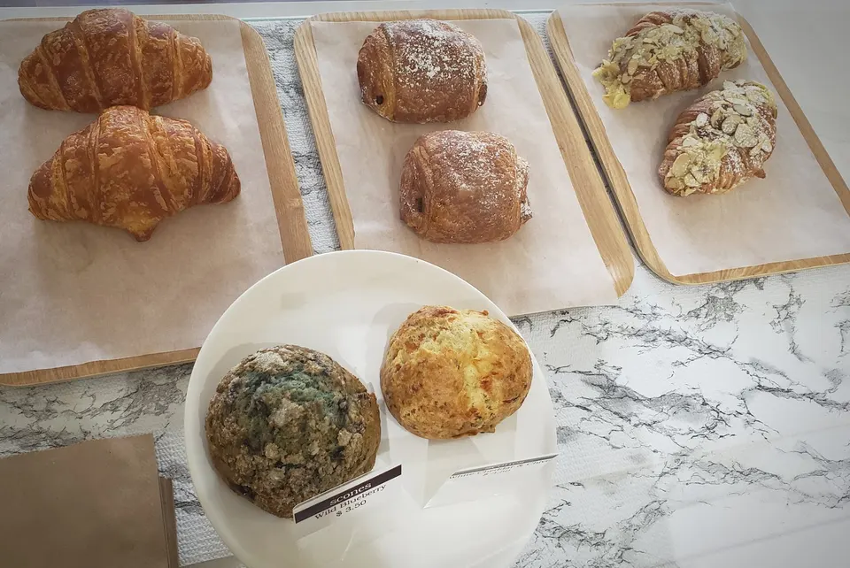 An assortment of freshly baked pastries displayed on wooden trays and a white plate, including croissants, pain au chocolat, almond croissants, and scones.