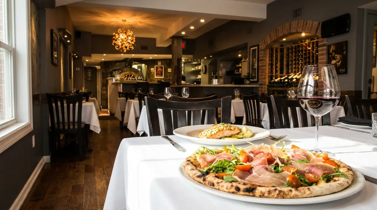 A freshly baked prosciutto and arugula pizza on a white plate next to a glass of red wine and a fish dish on a white tablecloth in a cozy restaurant dining room.