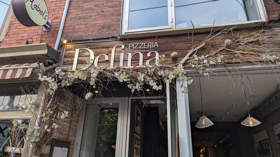 The storefront of Pizzeria Defina, featuring a rustic wooden sign with the restaurant name, decorated with dried branches and eucalyptus leaves above the entrance.