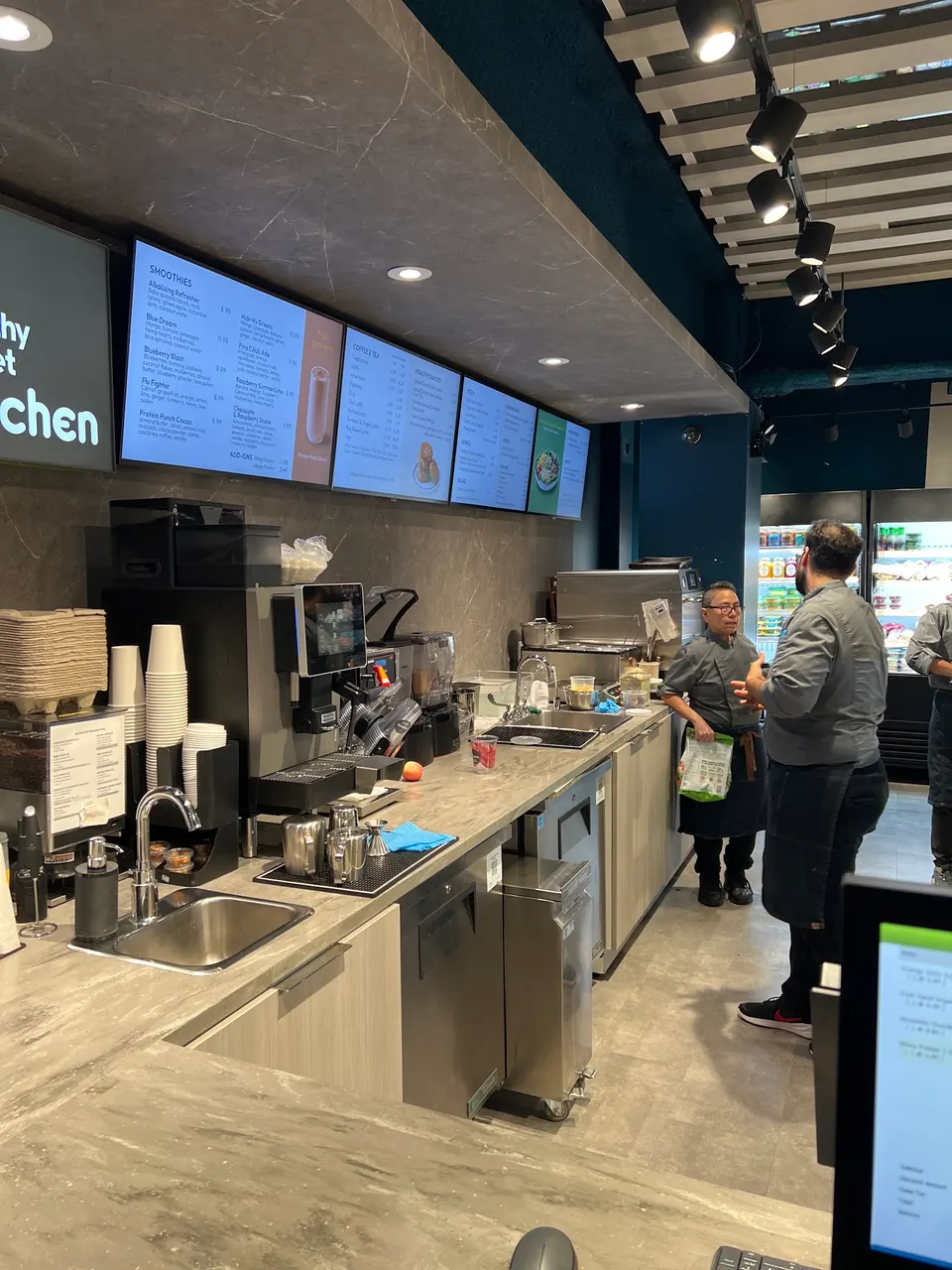 The counter area of Healthy Planet Kitchen, showing digital menu boards overhead, coffee and juice machines, stainless steel sinks, and two staff members in grey uniforms talking behind the counter.