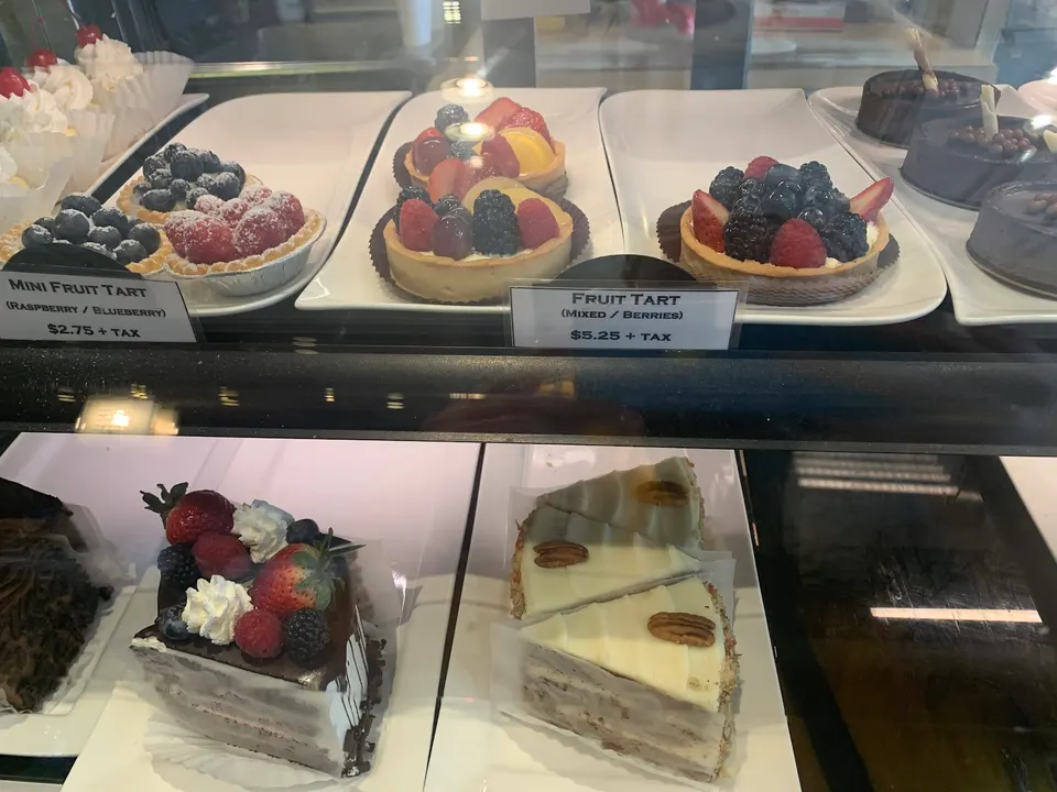 A bakery display case filled with various desserts including mini fruit tarts, larger fruit tarts with mixed berries, slices of chocolate berry cake, and slices of carrot cake with cream cheese frosting.