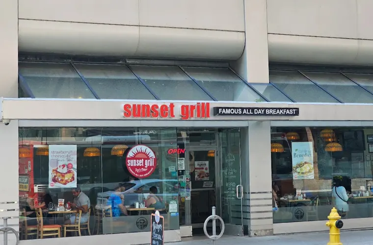Exterior storefront of Sunset Grill with red lettering on the sign, large glass windows showing diners inside, and a yellow fire hydrant on the sidewalk.