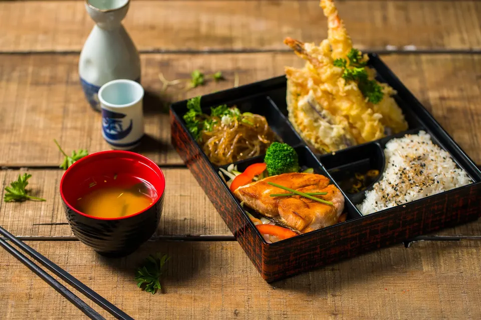 A Japanese bento box on a wooden table featuring tempura, grilled salmon, rice with sesame seeds, glass noodles, and a side of miso soup in a red and black bowl.