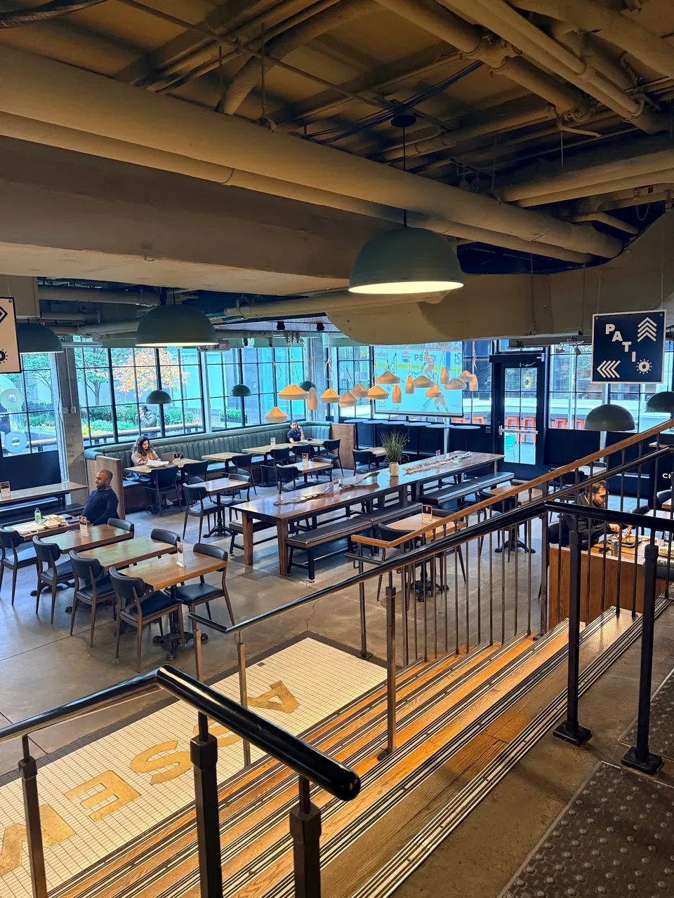 High-angle view of a spacious, modern restaurant dining area with industrial ceilings, long wooden communal tables, smaller dining sets, and stairs leading down to the main floor.