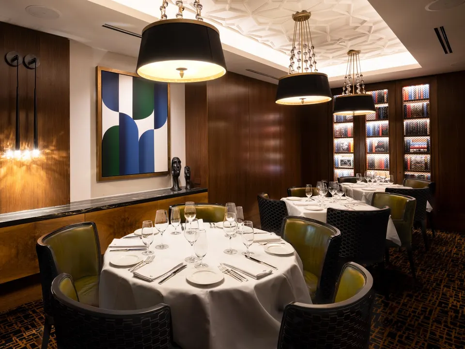 An elegant private dining room at Hy's Steakhouse & Cocktail Bar featuring round tables set with white tablecloths, fine glassware, and green leather chairs under large black drum pendant lights.