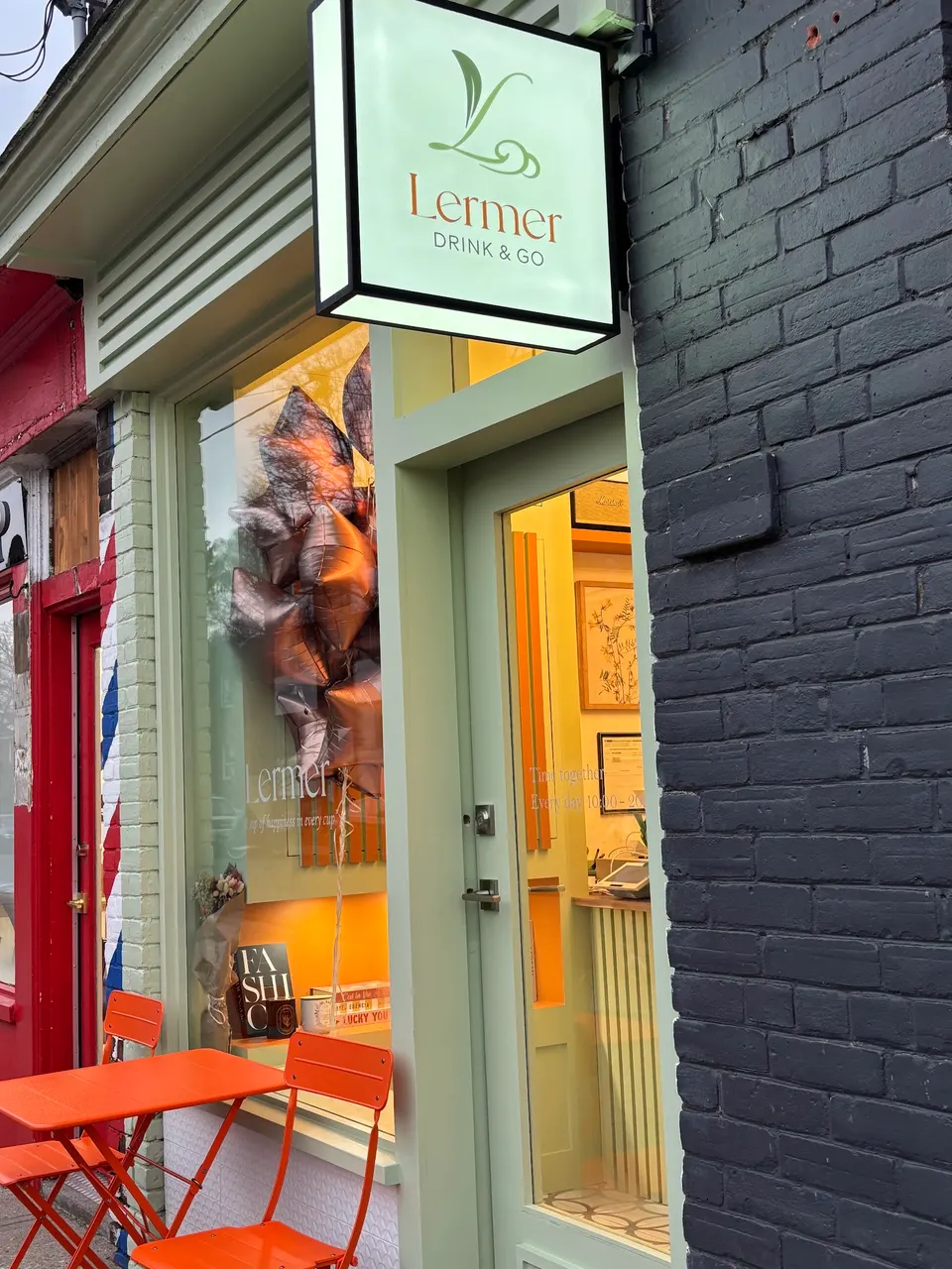 Exterior storefront of Lermer Cafe with a light green facade, a glowing square sign reading 'Lermer DRINK & GO', an orange outdoor table and chairs, and a glass door showing the warm interior.