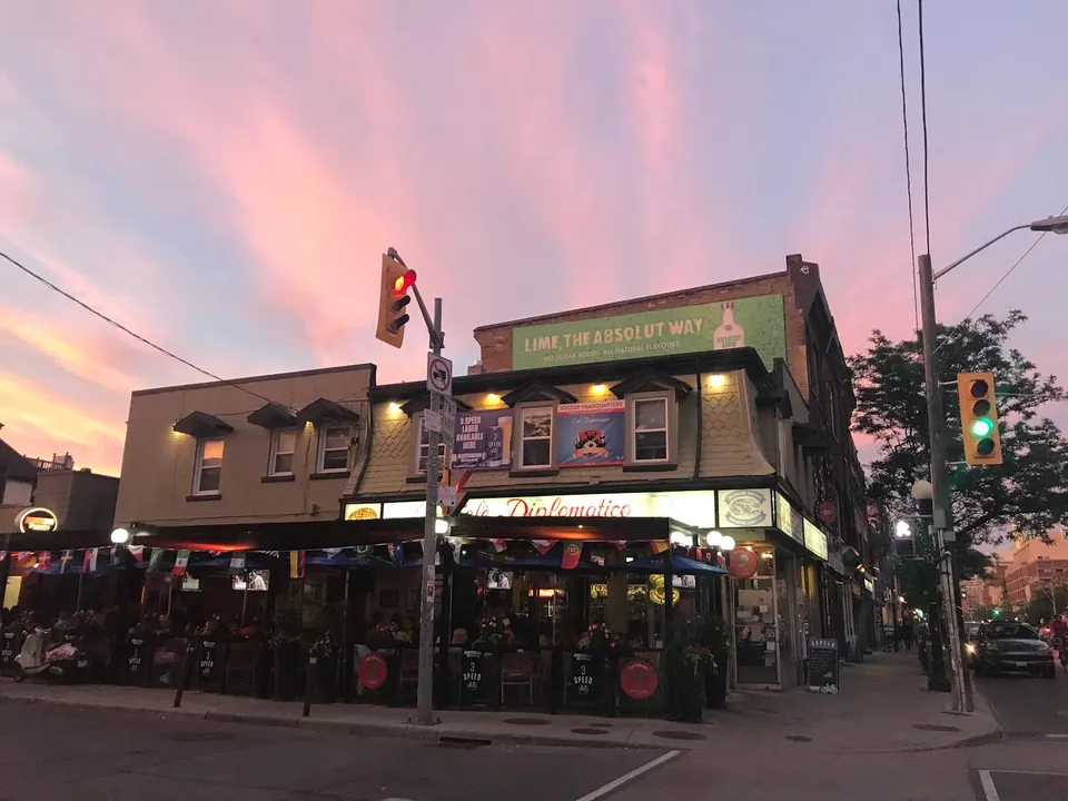 Exterior view of Cafe Diplomatico Restaurant & Pizzeria at dusk, featuring a lit-up storefront, patio seating under awnings, and a vibrant pink and purple sunset sky.