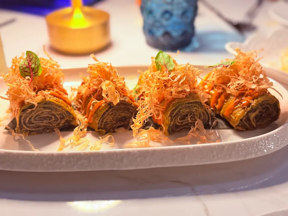 Four rolled savory crepes or pinwheels filled with a dark spiced mixture, drizzled with orange sauce, and topped with crispy shredded garnish and microgreens on a white platter.