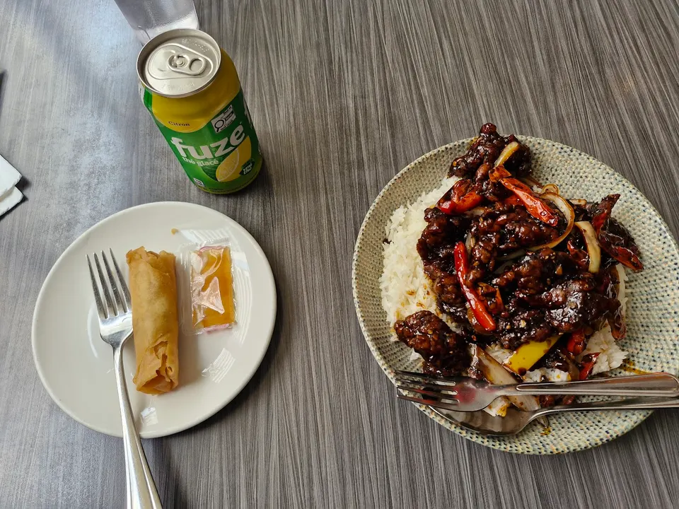 A table setting featuring a plate of stir-fried beef with peppers over white rice, a side plate with a spring roll and sweet sauce packet, and a can of Fuze iced tea.