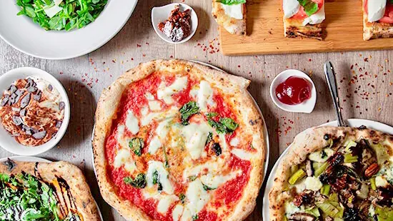 An overhead view of a wooden table filled with Italian dishes, featuring a classic Margherita pizza, a vegetable pizza, bruschetta on a wooden board, a green salad, and small bowls of sauces and toppings.