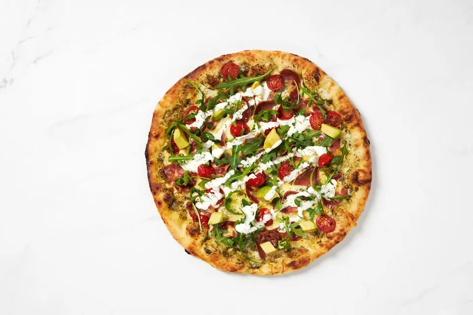 A whole wood-fired pizza on a white marble surface, topped with prosciutto, cherry tomatoes, avocado chunks, fresh arugula, and a drizzle of white cream sauce.