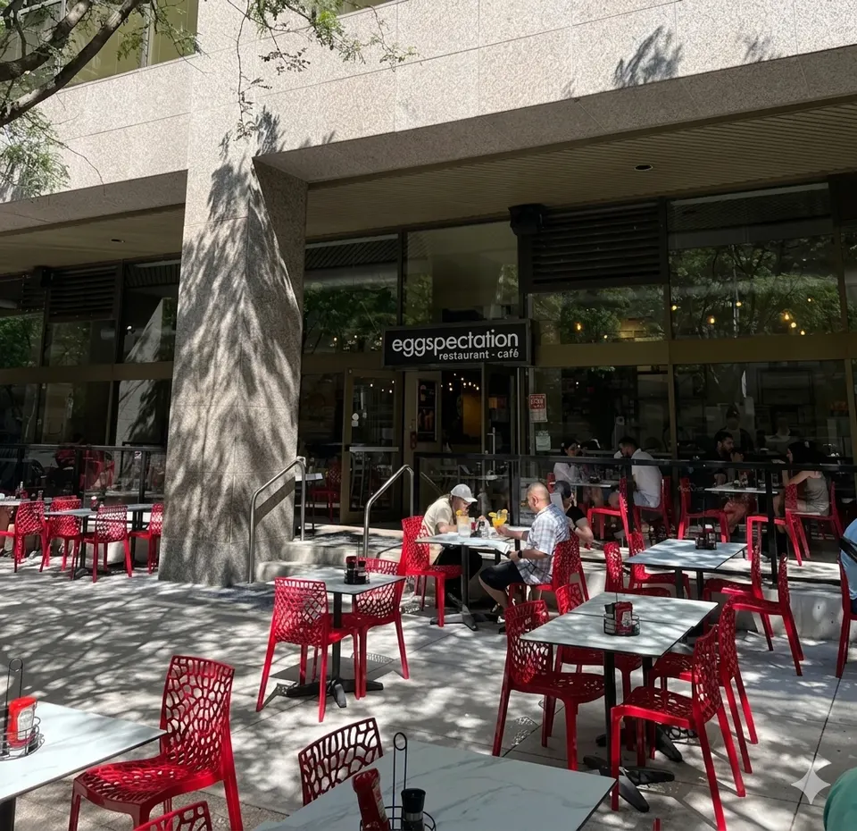 Outdoor patio of Eggspectation restaurant on Bay Street in Toronto, featuring red modern chairs, white tables, and patrons dining under a concrete overhang.