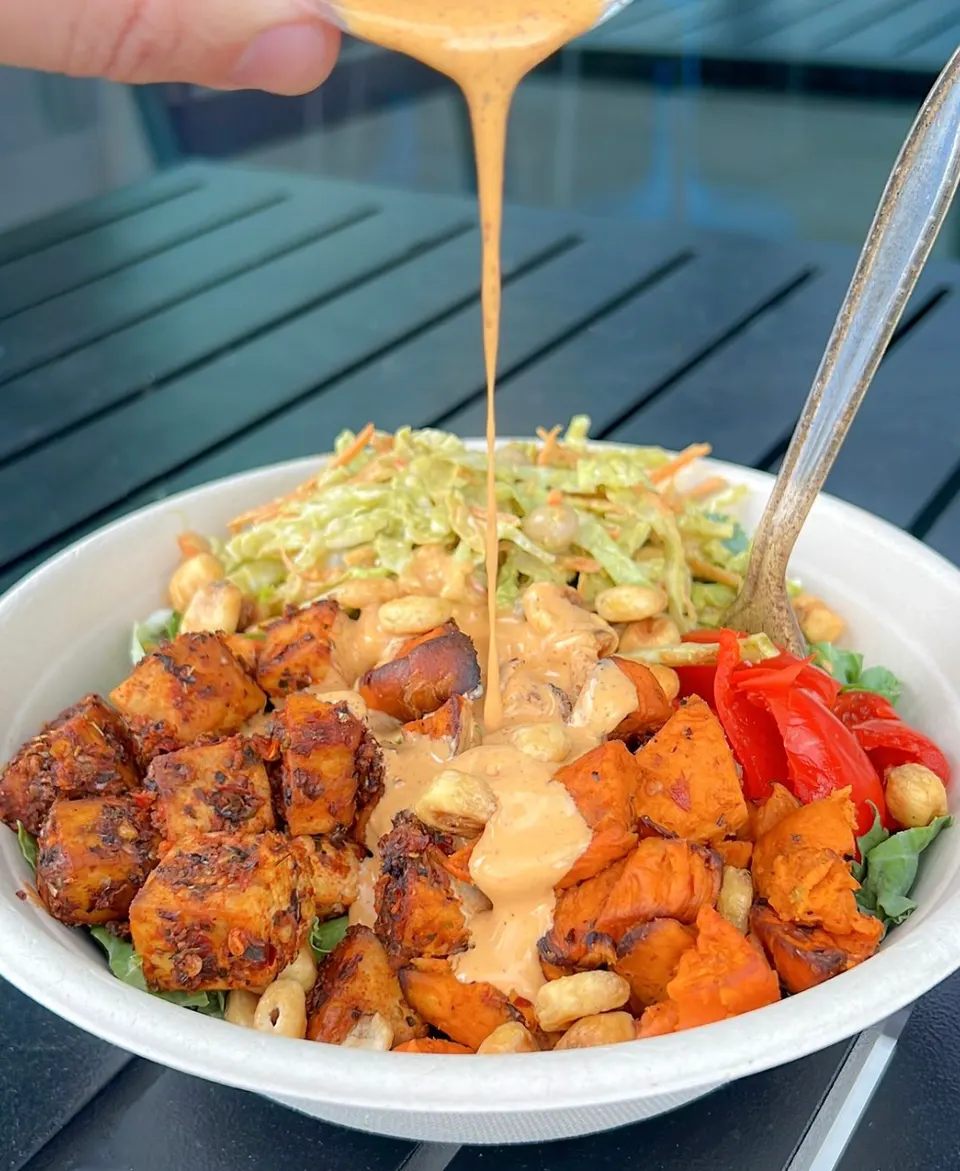 A healthy grain bowl filled with seasoned tofu cubes, roasted sweet potatoes, shredded cabbage slaw, red peppers, and crunchy nuts, being drizzled with a creamy orange dressing.