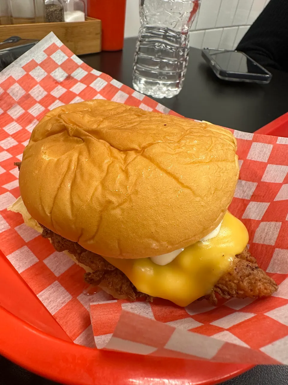 A close-up of a crispy chicken sandwich with melted yellow cheese and mayonnaise on a soft bun, served on red and white checkered paper in a red basket.