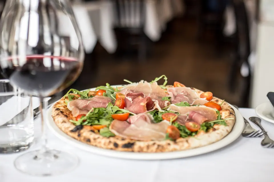 A freshly baked thin-crust pizza topped with prosciutto, fresh arugula, and cherry tomatoes on a white plate, with a glass of red wine in the foreground.