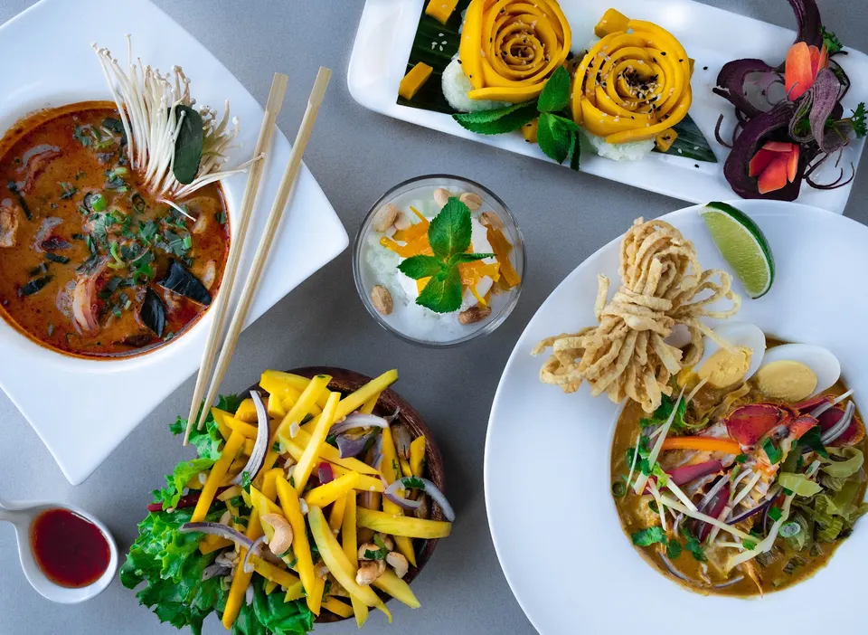 An overhead view of a table spread with Thai dishes, including a spicy soup with mushrooms, a mango salad, a noodle dish with crispy toppings, a dessert with mango roses, and a small bowl of coconut ice cream.