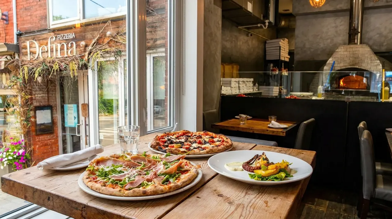 Two freshly baked wood-fired pizzas and a grilled octopus appetizer served on a rustic wooden table next to a large window at Pizzeria Defina, with the restaurant's exterior sign visible outside.