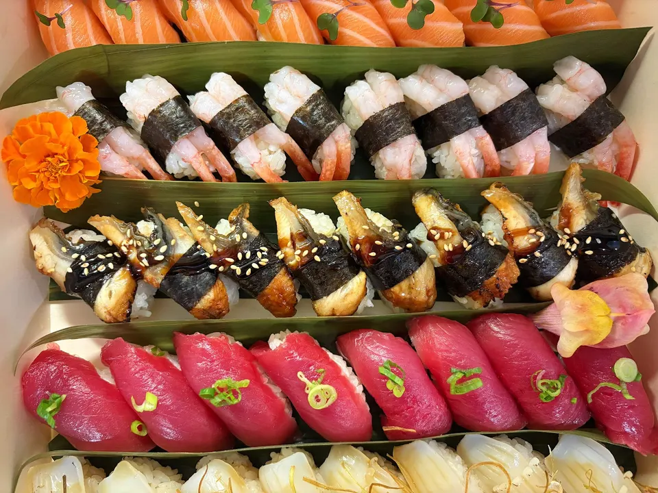 A close-up, top-down view of a premium sushi platter featuring rows of salmon, sweet shrimp, unagi (eel), tuna, and squid nigiri, separated by green leaves and garnished with edible flowers.