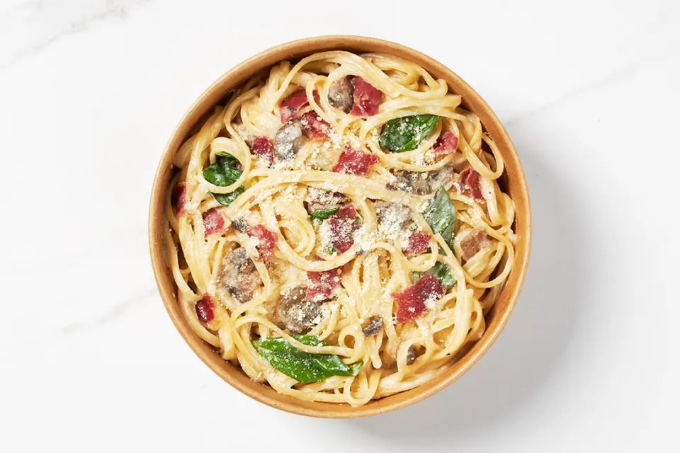 A top-down view of a round cardboard bowl filled with creamy fettuccine pasta, tossed with sliced mushrooms, spinach leaves, crispy bacon bits, and sprinkled with grated parmesan cheese.