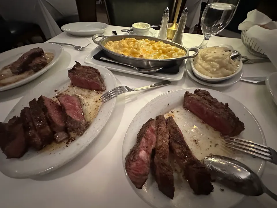 A table spread at Ruth's Chris Steak House featuring sliced medium-rare steaks on white plates, a metal dish of baked macaroni and cheese, mashed potatoes, and dining utensils.