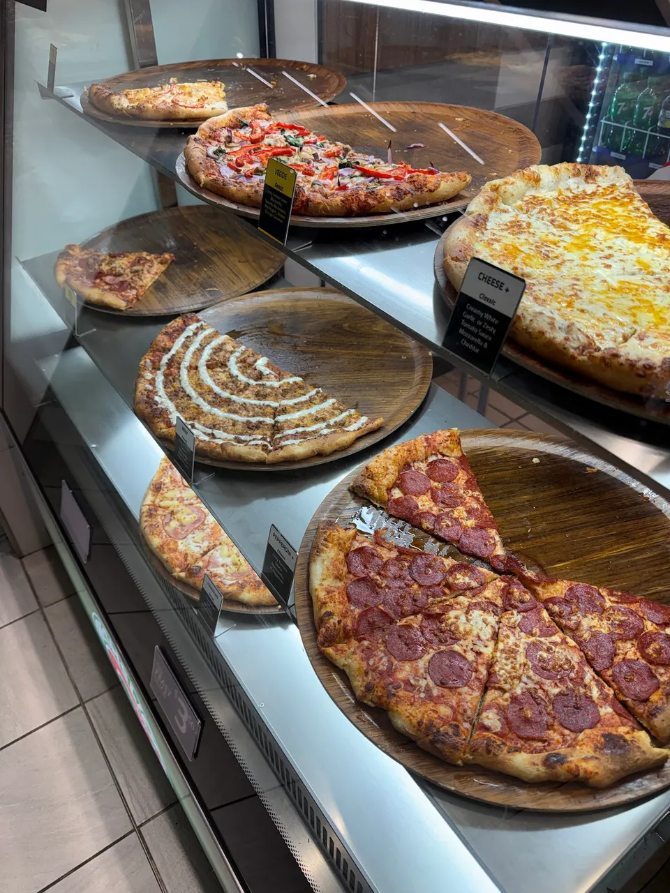 A glass display case filled with various freshly baked pizza slices on wooden serving trays, including pepperoni, cheese, veggie, and white sauce options.