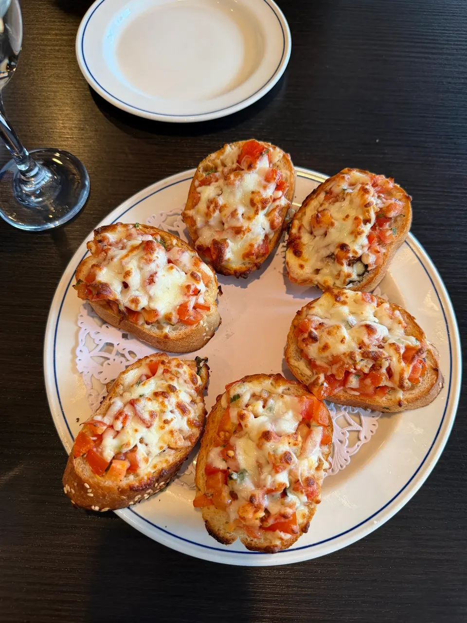 Six pieces of toasted bruschetta topped with diced tomatoes and melted mozzarella cheese, arranged in a circle on a white plate with a blue rim.