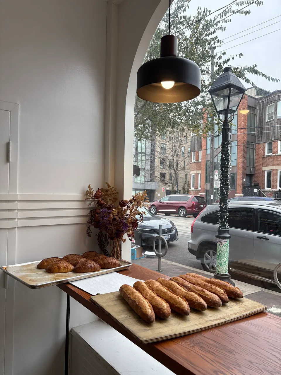Freshly baked baguettes and pastries rest on wooden boards and trays on a counter inside Bricolage Bakery, looking out through a large arched window onto a city street with parked cars and a vintage street lamp.