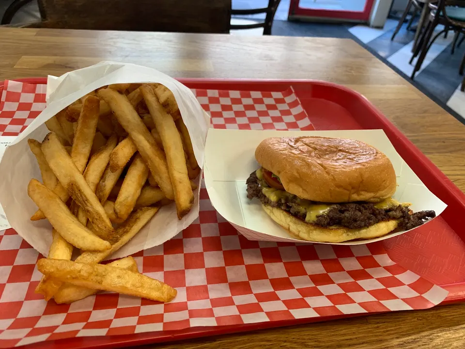 A smash burger with melted cheese on a bun sits in a paper tray next to a large portion of golden french fries overflowing from a white paper bag on a red and white checkered liner.